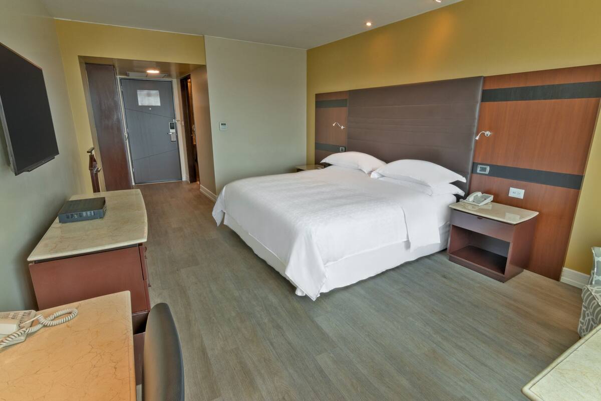 A hotel room with a large bed, a desk, a chair, a bedside table, and a wardrobe.