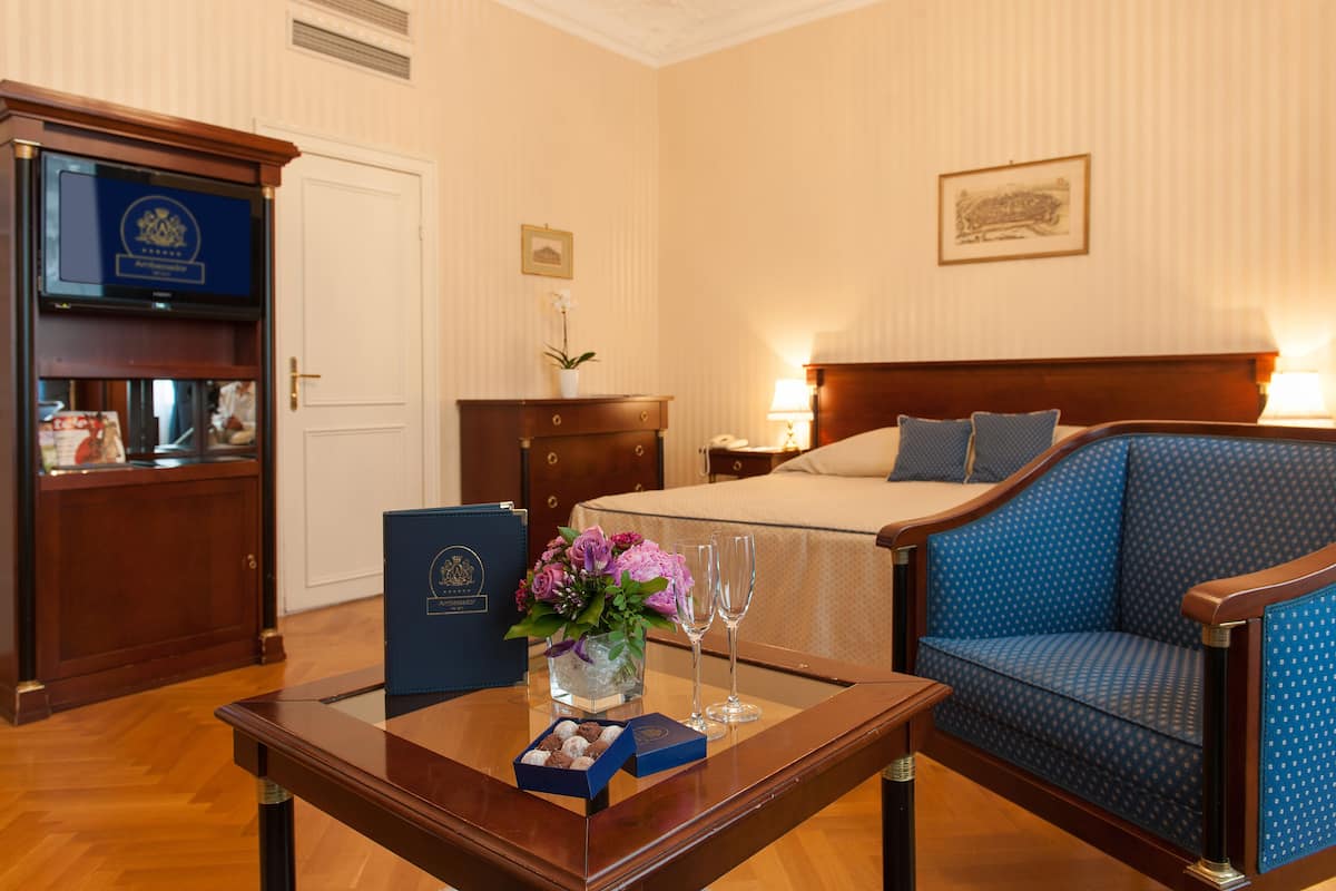 Quarto de hotel com uma cama, uma poltrona estofada azul, uma escrivaninha de madeira com um vaso de flores, uma caixa azul e duas taças de champanhe.