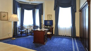 A hotel room with a large desk, two chairs, a chandelier, and a television.