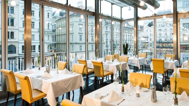 A restaurant with tables set for dining, large windows offering city views, and yellow chairs.