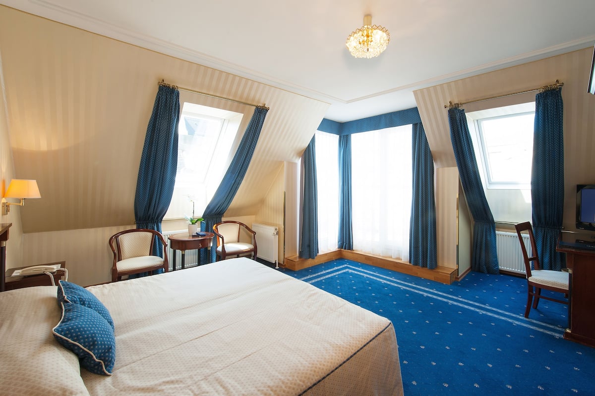 Quarto de hotel com uma cama grande, duas poltronas, uma mesinha, uma secretária com computador e uma janela com cortinas.