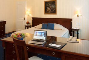 A hotel room with a bed, a desk with a laptop, a telephone, and a small table with a fruit bowl.