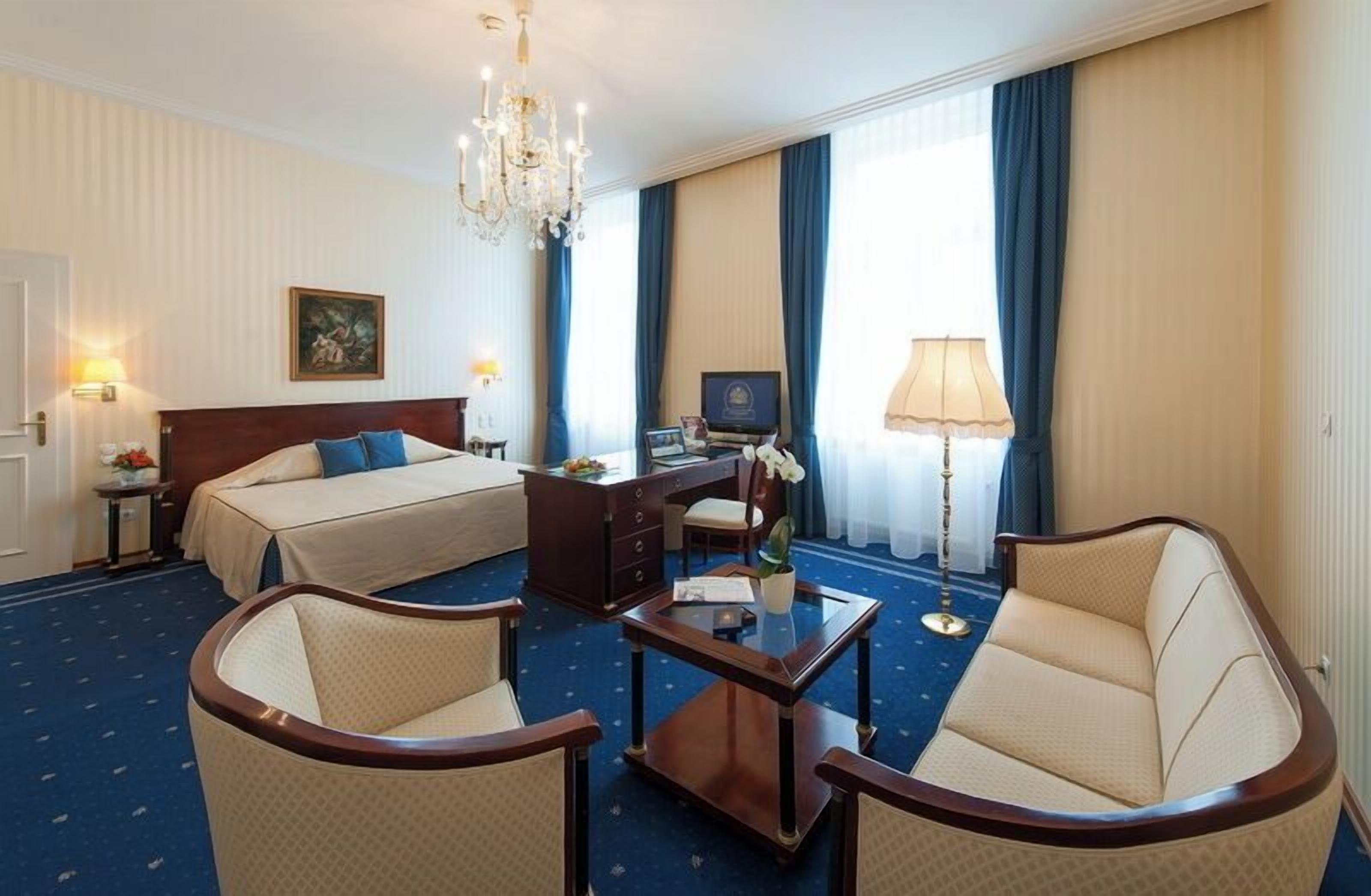 Une chambre d’hôtel avec un lit, deux fauteuils, un bureau, un lustre et une grande fenêtre avec des rideaux.
