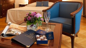 A neatly arranged hotel room with a bed, a blue upholstered chair, a glass coffee table with a magazine, a vase of flowers, a glass of champagne, and two boxes of chocolates.