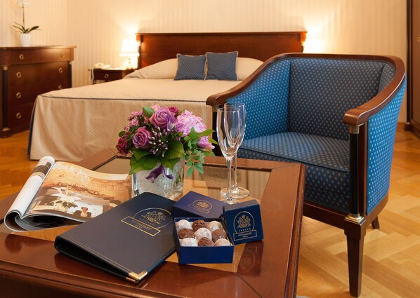A neatly arranged hotel room with a bed, a blue upholstered chair, a glass coffee table with a magazine, a vase of flowers, a glass of champagne, and two boxes of chocolates.