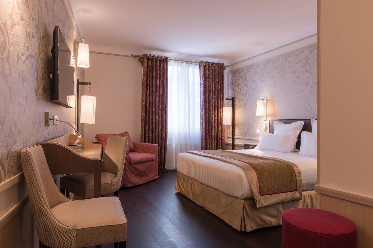 A hotel room with a bed, a desk with a chair, a red ottoman, and a TV mounted on the wall.
