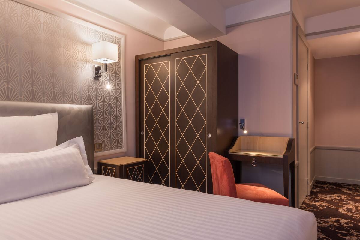 A hotel room with a large bed, a wardrobe with a geometric pattern, a small desk with a lamp, and a bathroom with a mirror and sink.