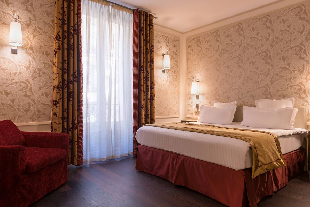 A hotel room with a large bed, red curtains, patterned wallpaper, and two wall-mounted lamps.
