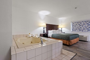 A hotel room with a large bathtub, a bed with a green and orange bedspread, two bedside lamps, and a wall-mounted air conditioner.
