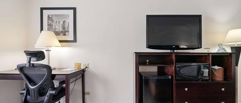 Habitación de hotel con escritorio, una silla de oficina negra, un televisor sobre un mueble de madera, un microondas y una lámpara.