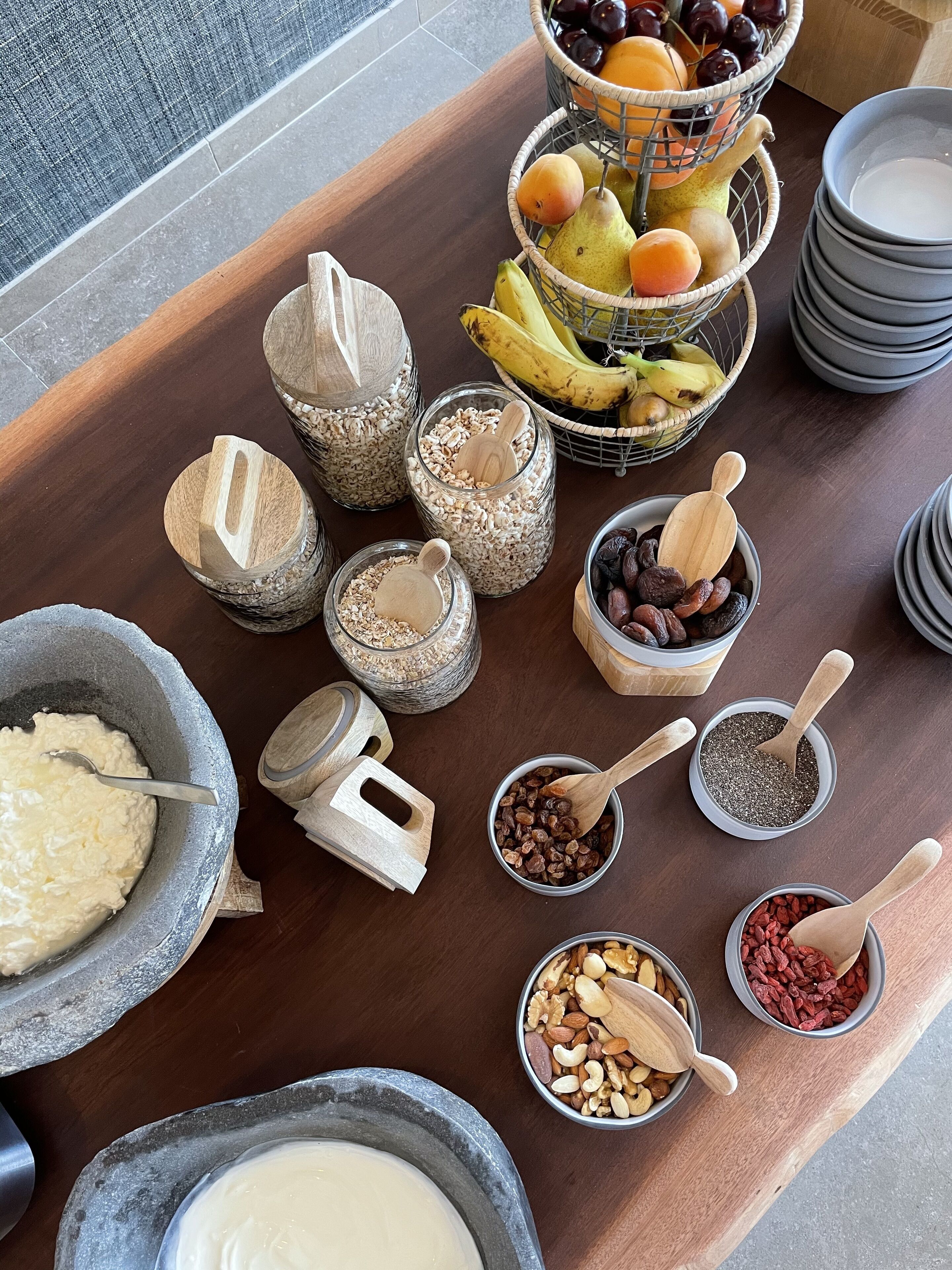 A wooden table with various food items and utensils, including bowls of grains, nuts, and dried fruits, as well as a fruit basket and stacked plates.