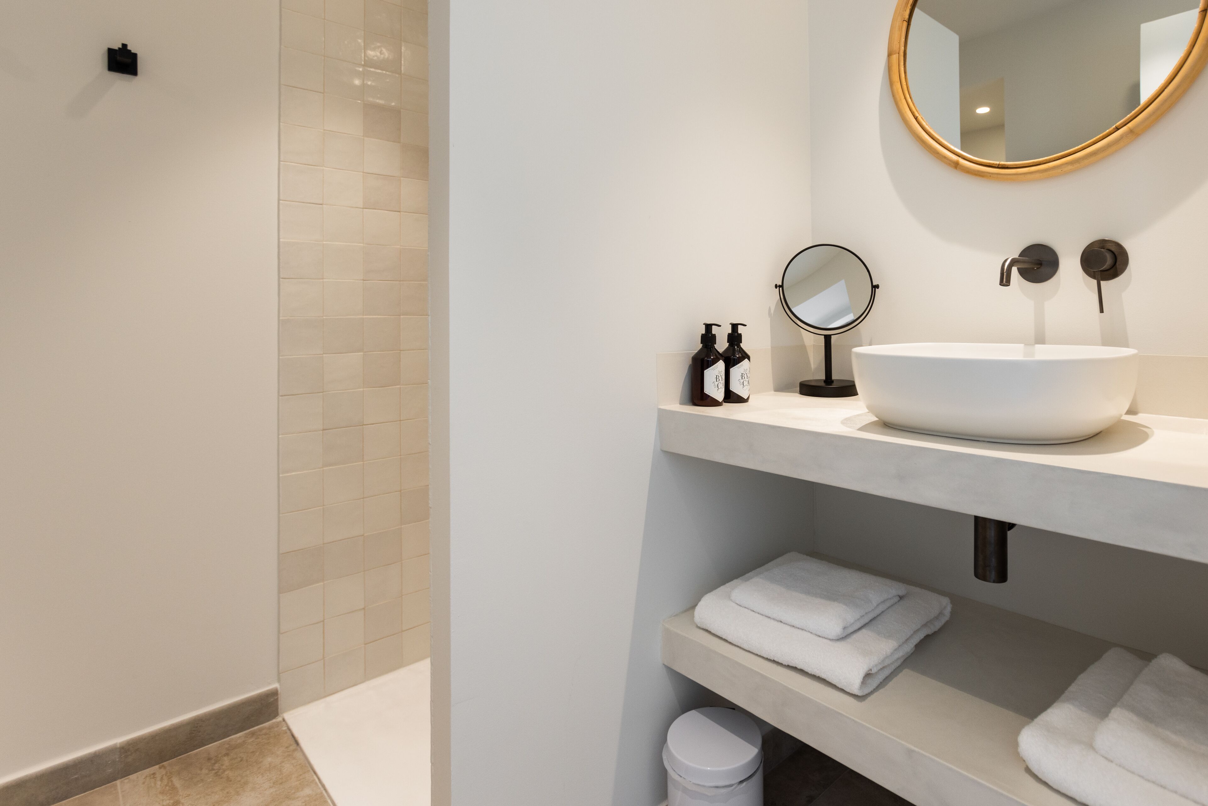 Een moderne badkamer met een groot wit wastafelmeubel, een ronde spiegel en netjes opgevouwen witte handdoeken op planken.