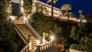 A grand staircase leading to a building with multiple balconies, surrounded by lush greenery and palm trees. - Le Mirage (Bormes-Les-Mimosas)