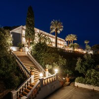 Una imponente escalera conduce a un edificio con varios balcones, rodeado de exuberante follaje y palmeras.