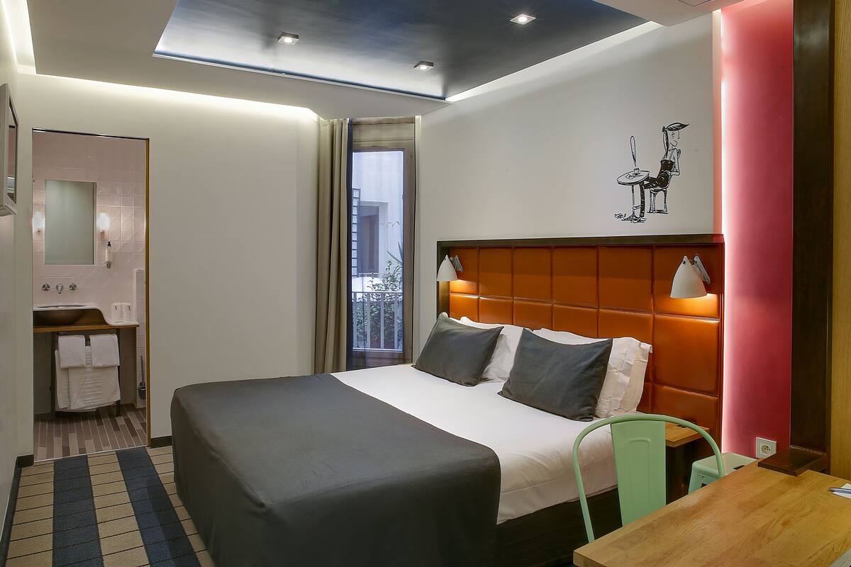 Una habitación de hotel moderna con una cama grande, cabecera de madera, una mesita, una silla verde y un baño con lavamanos y espejo.