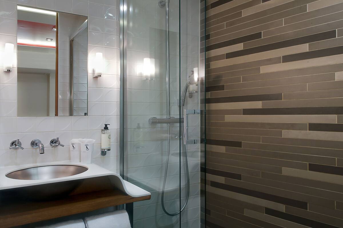 Un baño moderno con ducha de vidrio, tocador de madera con lavamanos y pared de azulejos con diseño de ladrillo.