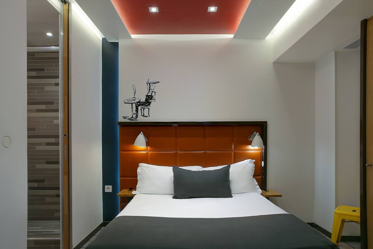 Una habitación de hotel moderna con una cama grande, cabecera con iluminación integrada, una mesita pequeña y una obra de arte colgada en la pared.