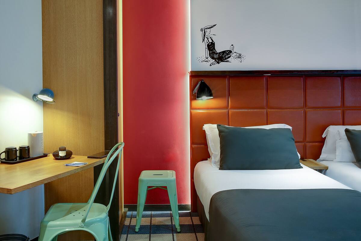 Habitación de hotel con un escritorio de madera, un taburete pequeño, una cama con dos almohadas, una mesita de noche y una lámpara de pared.
