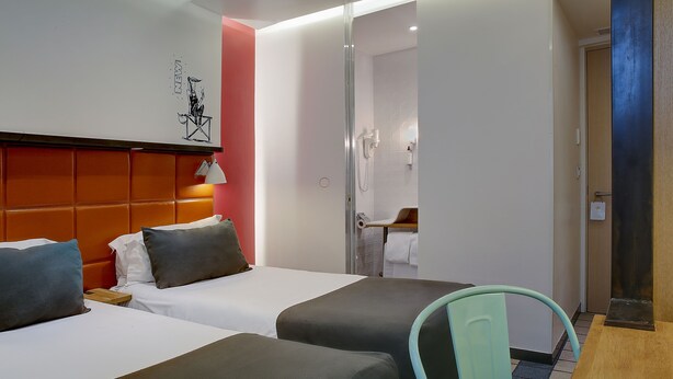 Une chambre d’hôtel avec deux lits, une tête de lit en bois, un miroir, une salle de bain avec douche et une petite table avec une chaise.