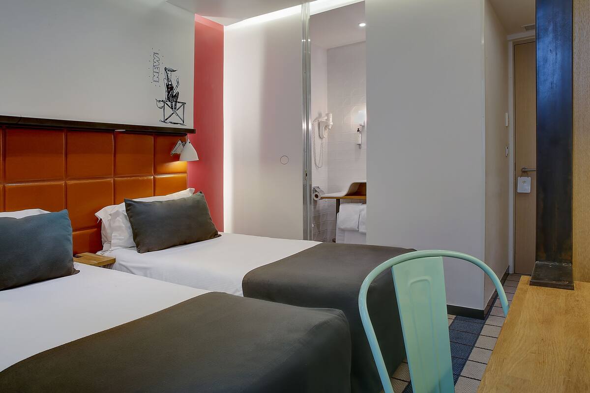 Habitación de hotel con dos camas, cabecero de madera, espejo, baño con ducha y una mesita con silla.