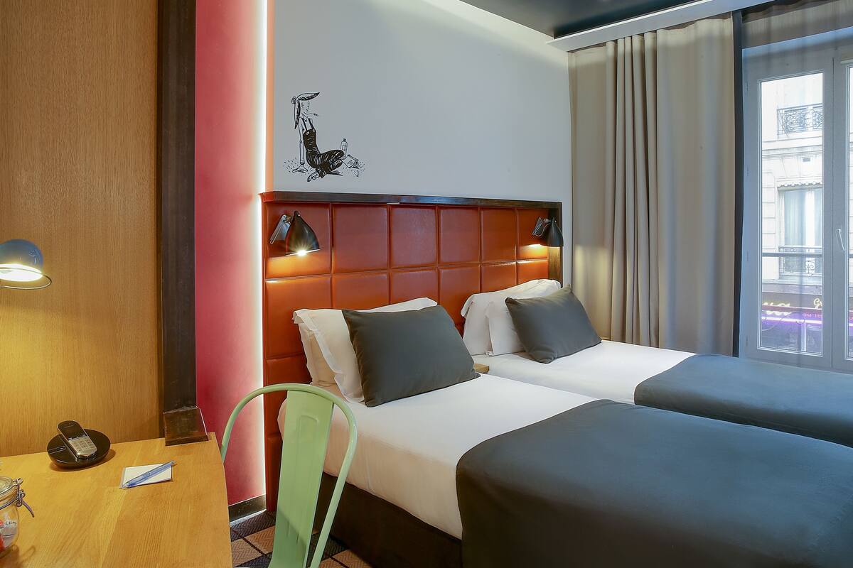 Habitación de hotel con dos camas, un escritorio de madera, una silla verde y una obra de arte colgada en la pared.