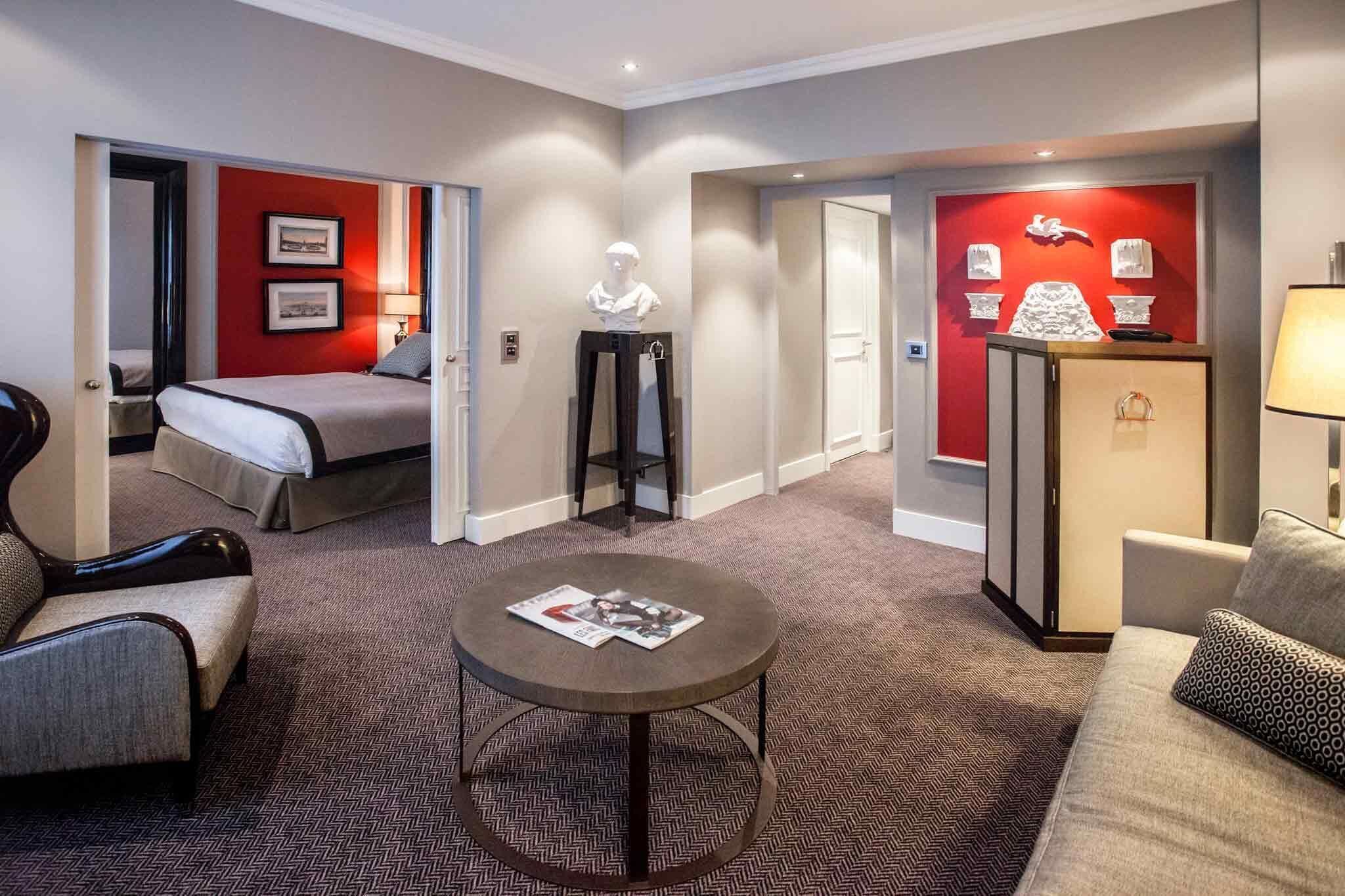 A hotel room with a bed, a round table, a sofa, a cabinet, and a red accent wall.