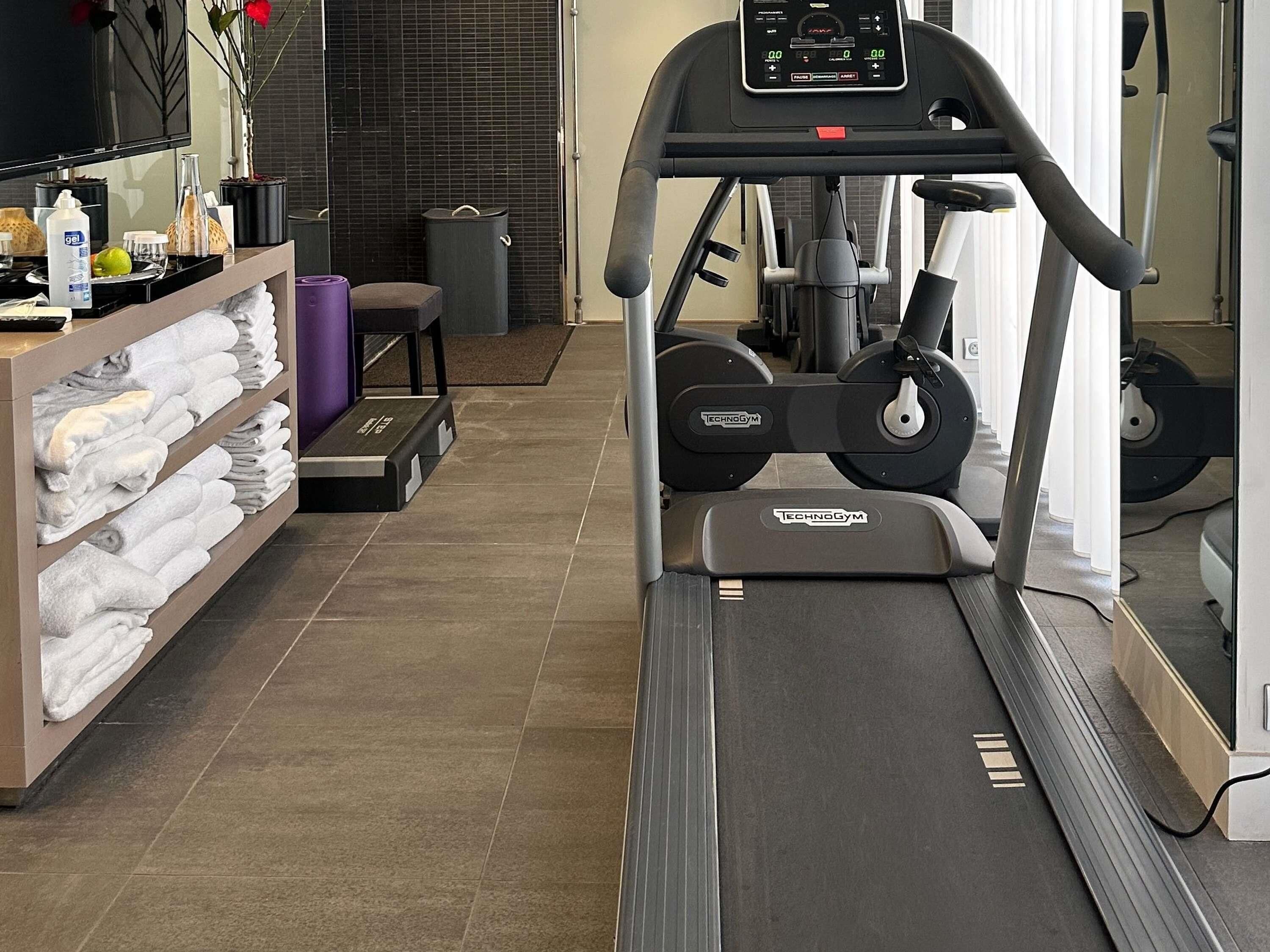 Un gimnasio con cinta de correr, equipo de ejercicio, un perchero para toallas y un cesto de basura.