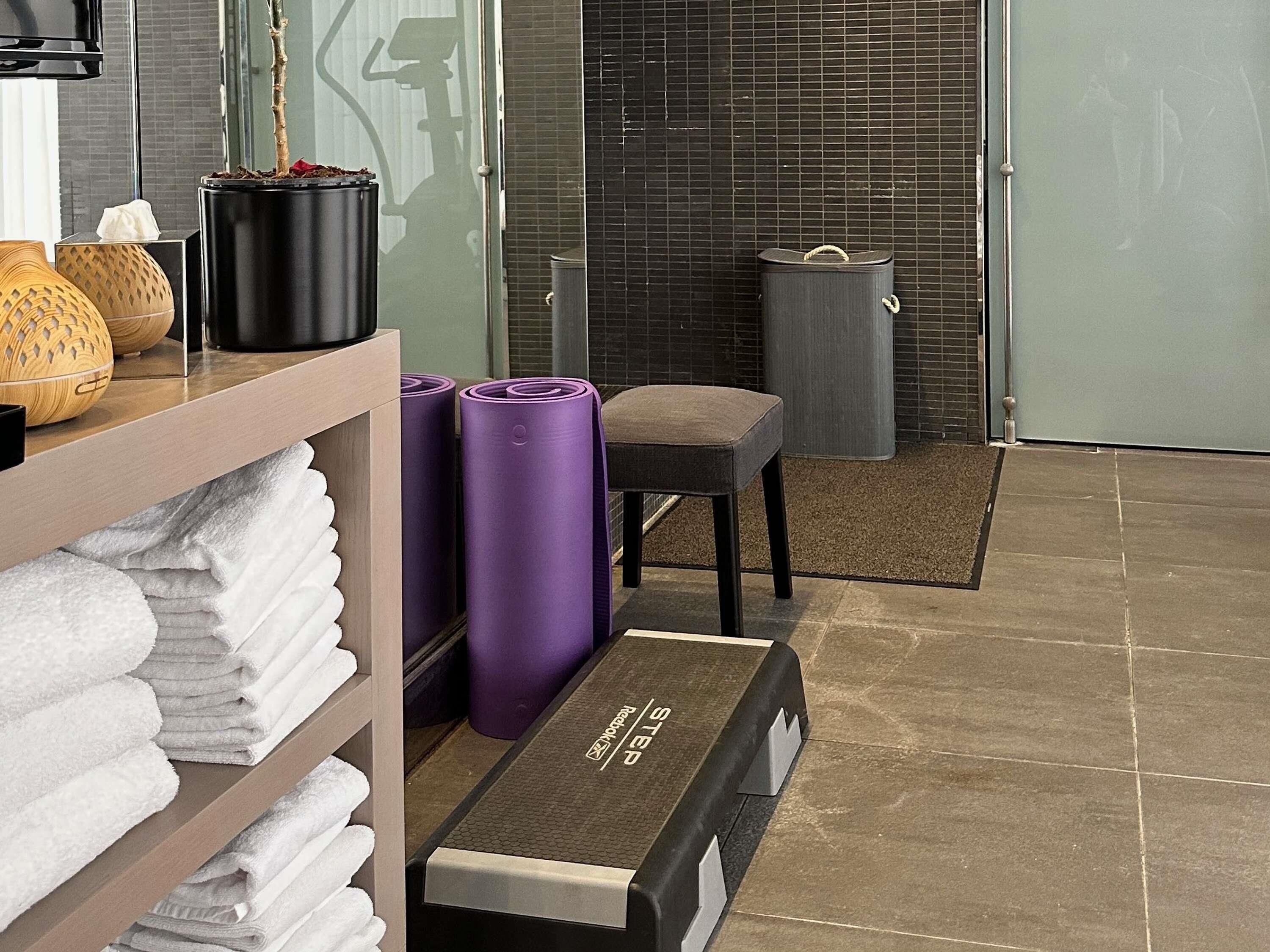 A hotel room with a wooden shelf holding towels, a black trash bin, a grey chair, and a yoga mat.