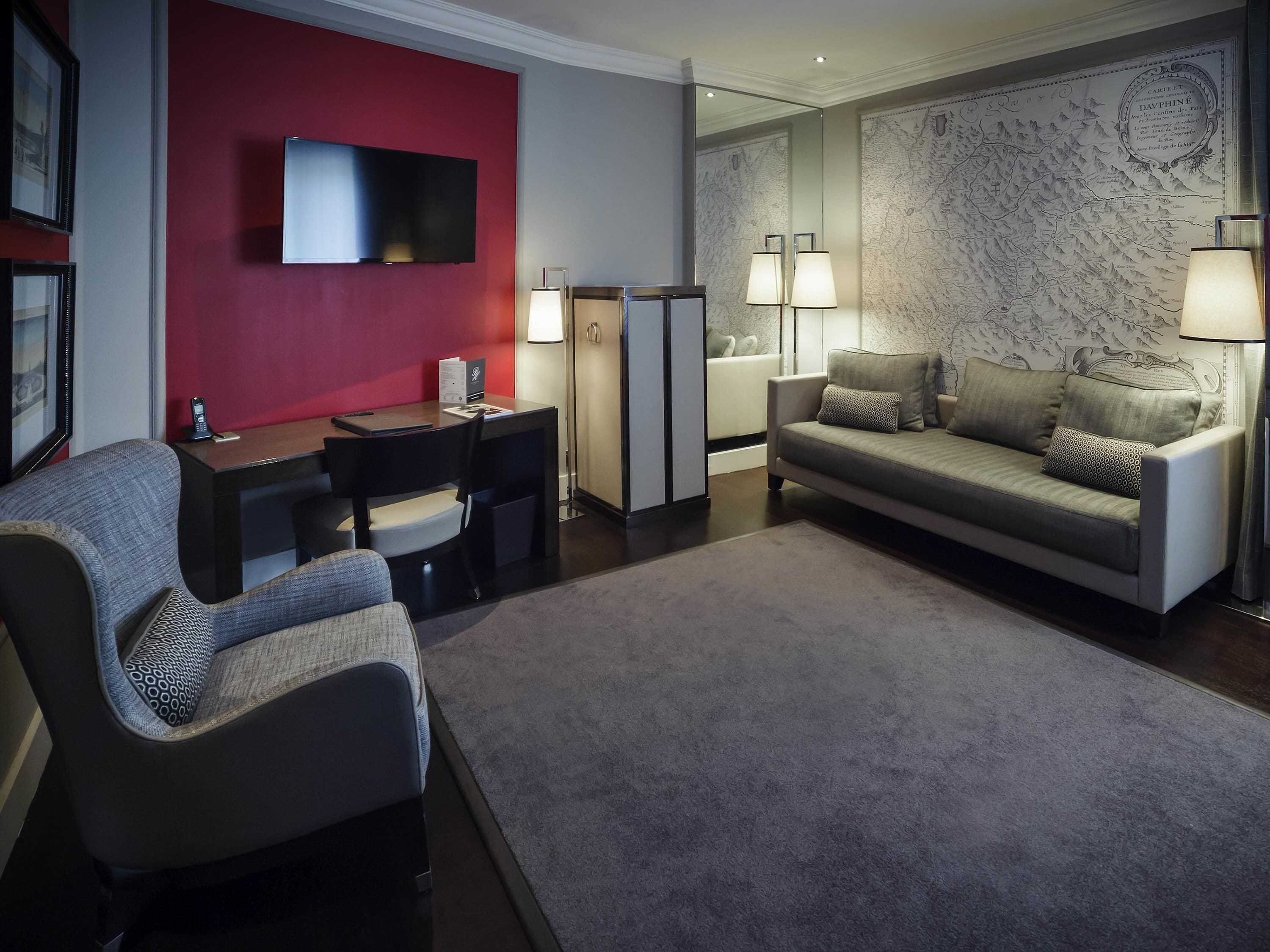 A modern hotel room with a red accent wall, a flat-screen TV, a desk with a chair, a sofa, and two floor lamps.