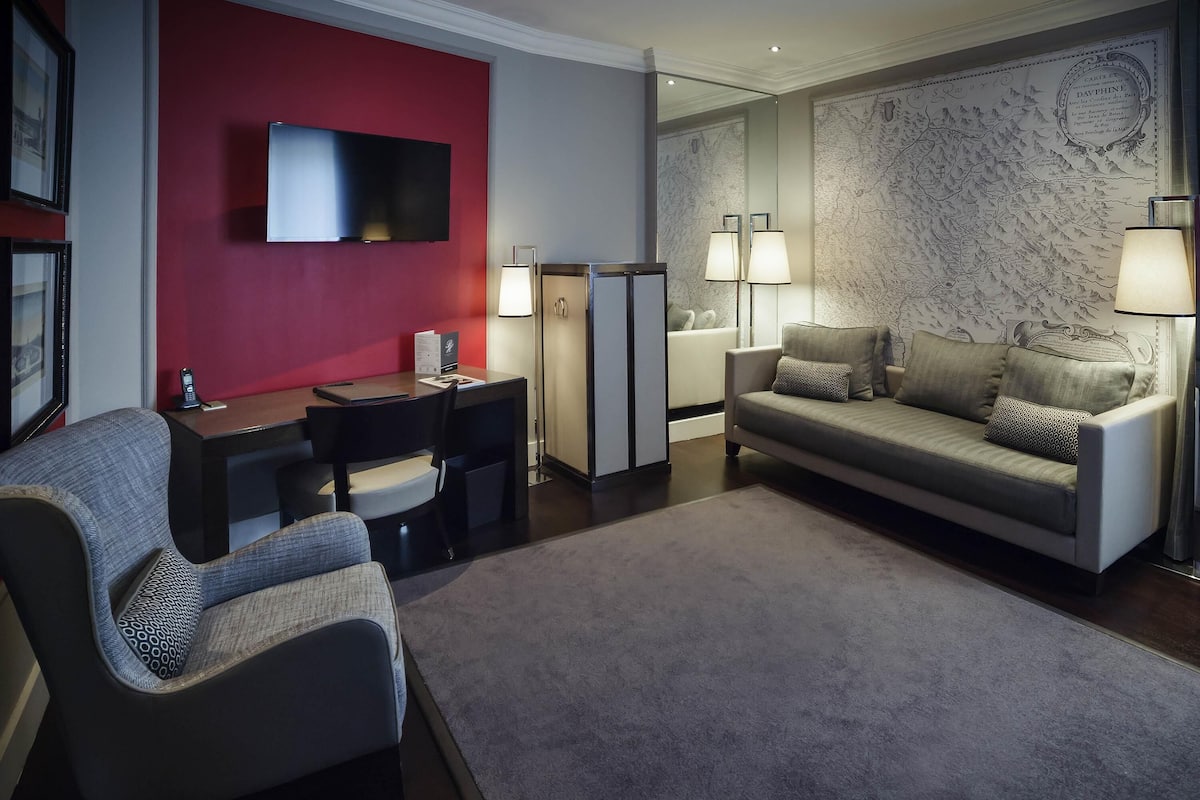 Une chambre d’hôtel moderne avec un mur d’accent rouge, un téléviseur à écran plat, un bureau avec une chaise, un canapé et deux lampadaires.