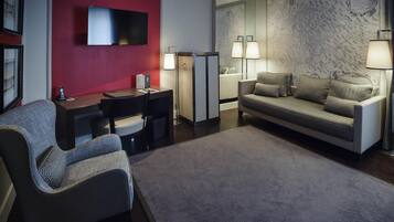 A modern hotel room with a red accent wall, a flat-screen TV, a desk with a chair, a sofa, and two floor lamps.