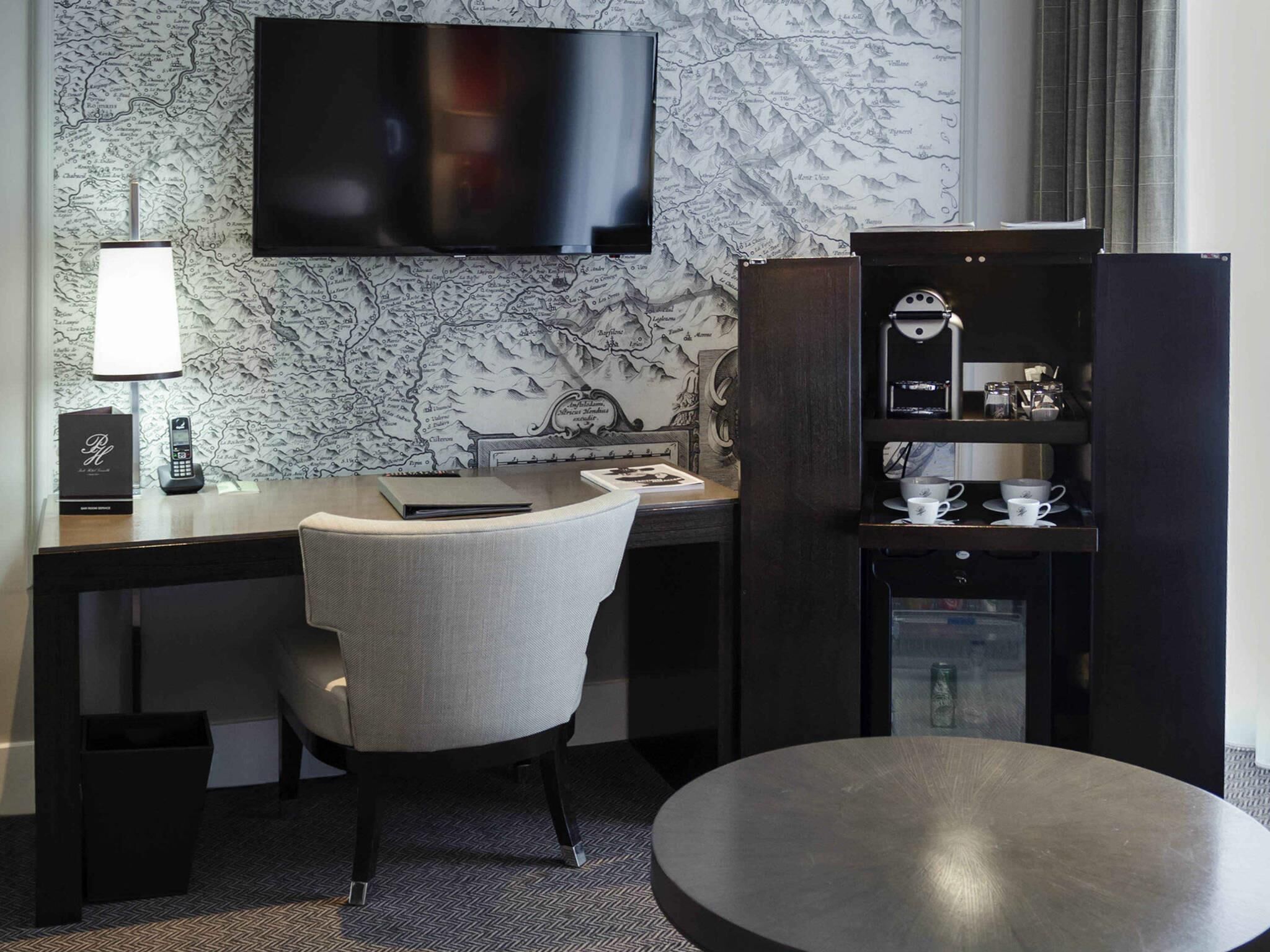 A hotel room with a flat-screen TV, a desk with a lamp, a chair, a bookshelf, and a coffee machine.