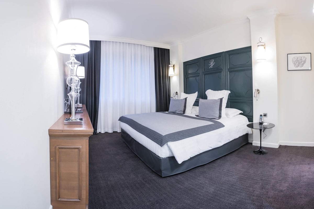 Una habitación de hotel con una cama grande, una mesita de noche con lámpara, un escritorio con teléfono y una lámpara de pared.