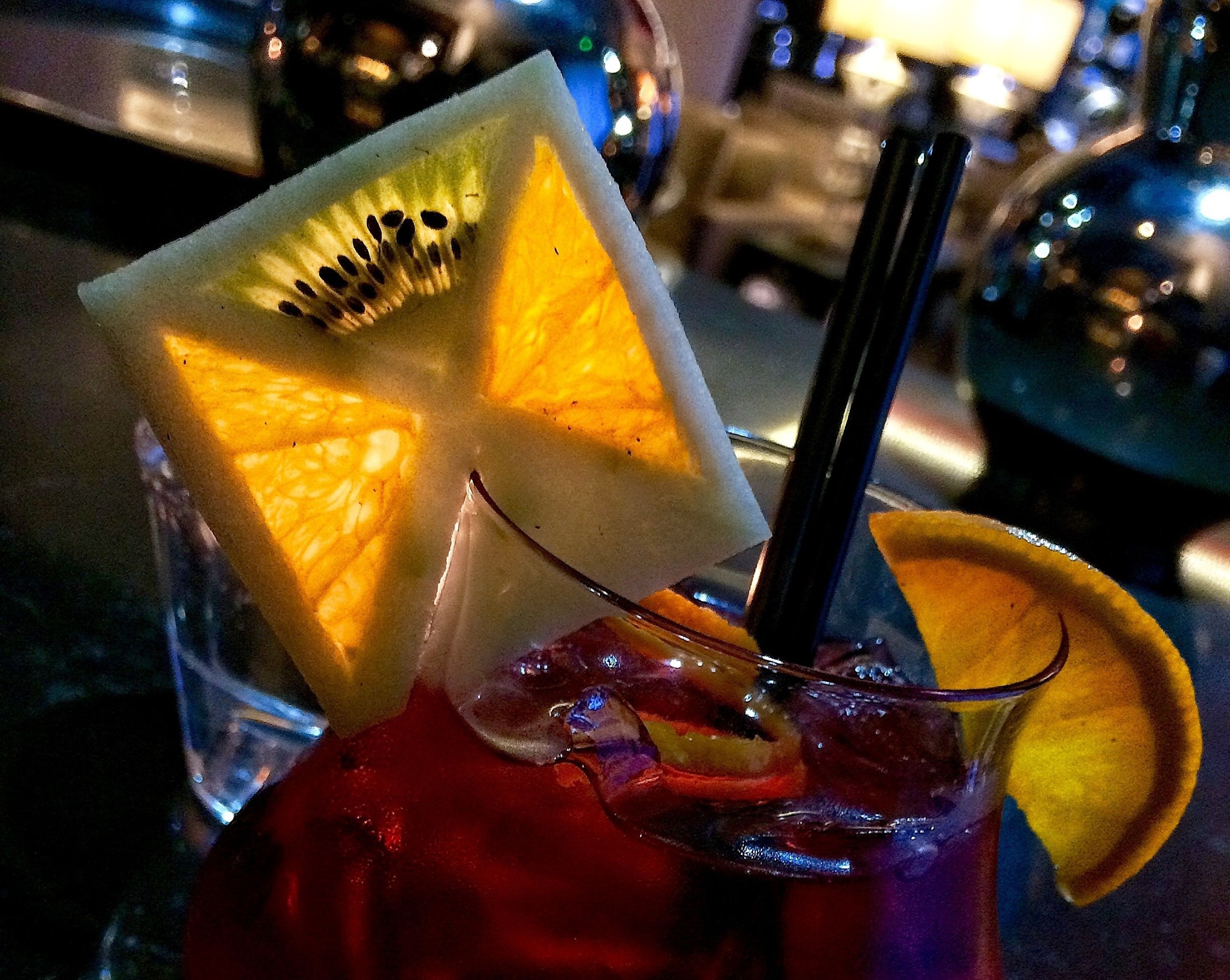 A cocktail with a slice of kiwi and an orange slice, a straw, and ice cubes in a glass.