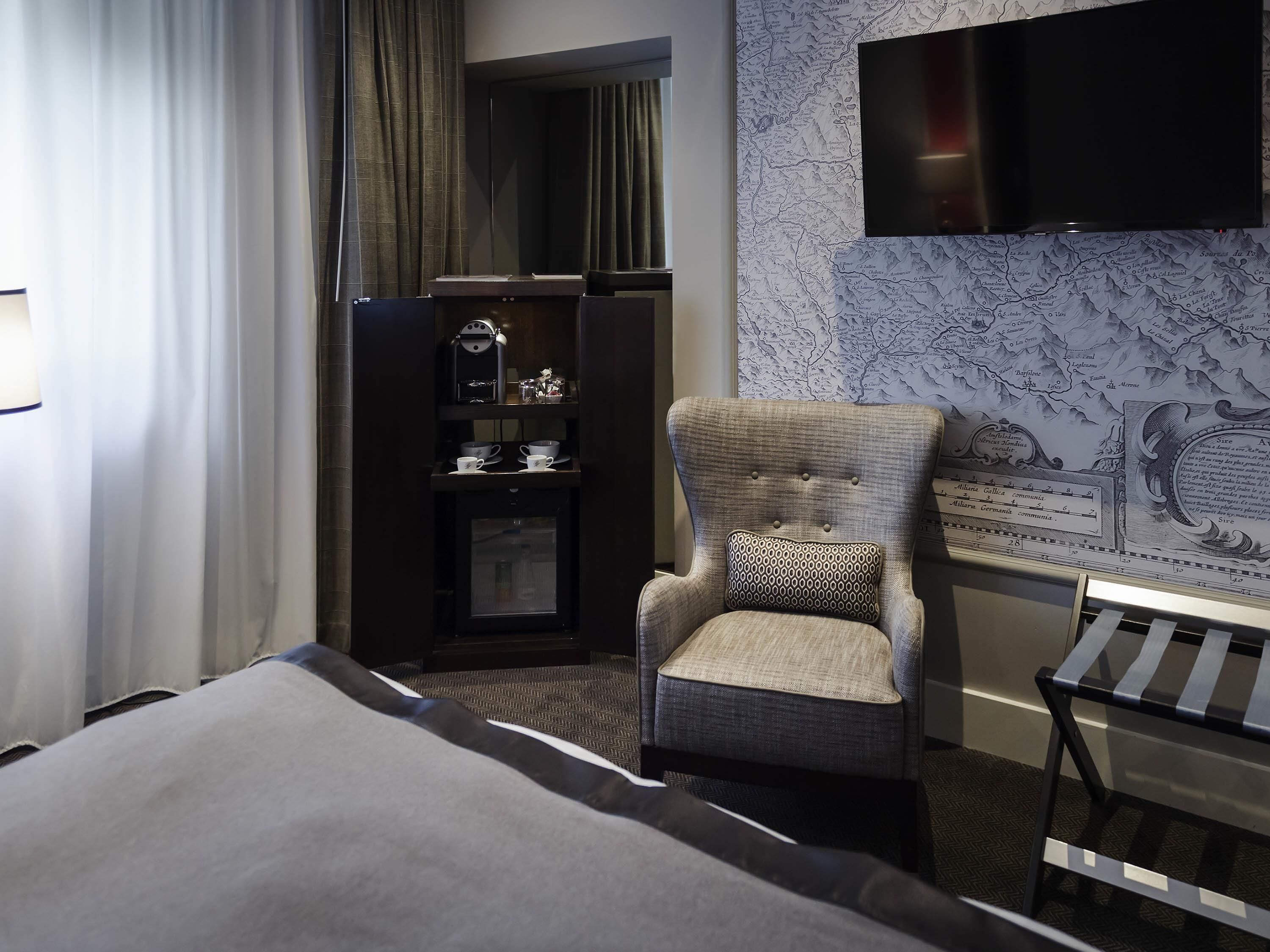 A hotel room with a bed, a chair with a patterned cushion, a small table, a shelf with cups and a coffee machine, and a wall-mounted TV.