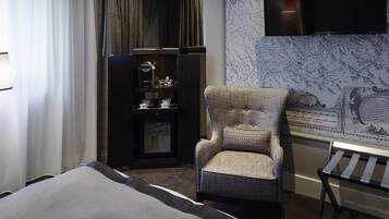 A hotel room with a bed, a chair with a patterned cushion, a small table, a shelf with cups and a coffee machine, and a wall-mounted TV.