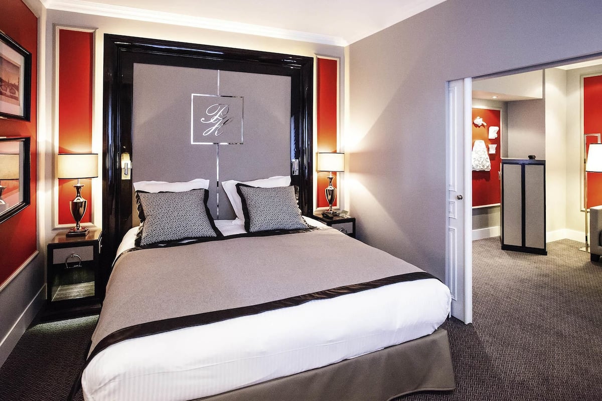 Una habitación de hotel con una cama grande, dos mesitas de noche con lámparas, un cabecero con un logotipo, una puerta al baño y una pared decorativa roja.
