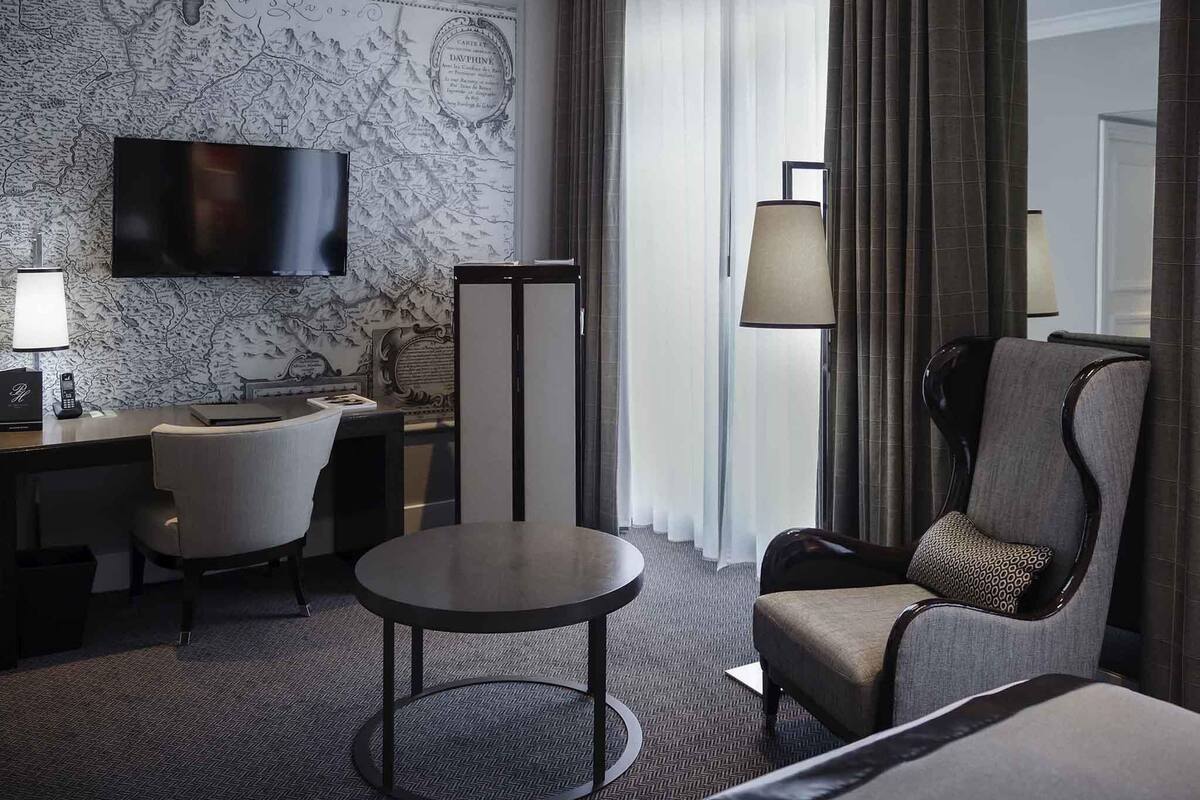 Een hotelkamer met een flatscreen televisie, een bureau met lamp, een ronde tafel, een bank en een groot raam met gordijnen.