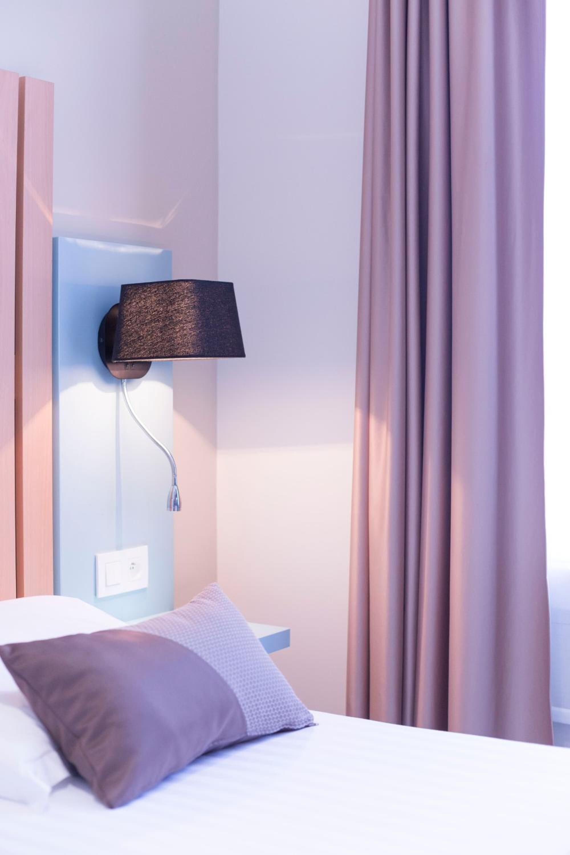 A hotel room with a bed, a wall-mounted lamp, and a window with curtains.