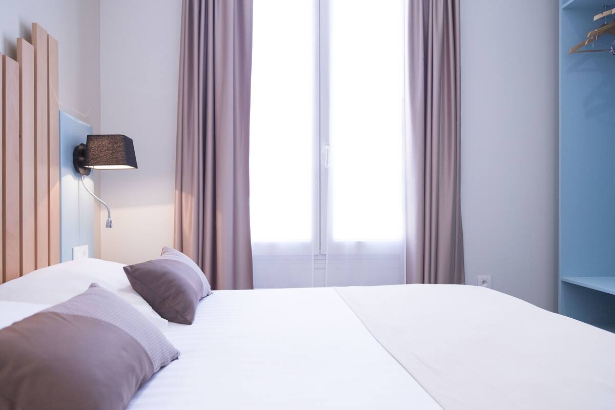 A hotel room with a large bed, a wall-mounted lamp, and a window with curtains.