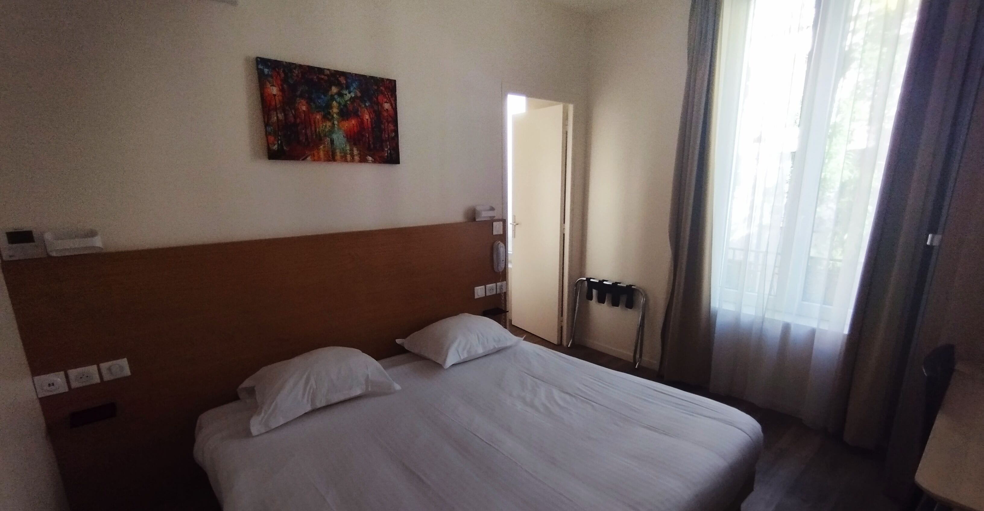 Una habitación de hotel con una cama grande, un cabecero, un cuadro en la pared, una ventana con cortinas y una puerta que da a otra habitación.