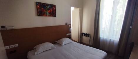 Una habitación de hotel con una cama grande, un cabecero, un cuadro en la pared, una ventana con cortinas y una puerta que da a otra habitación.