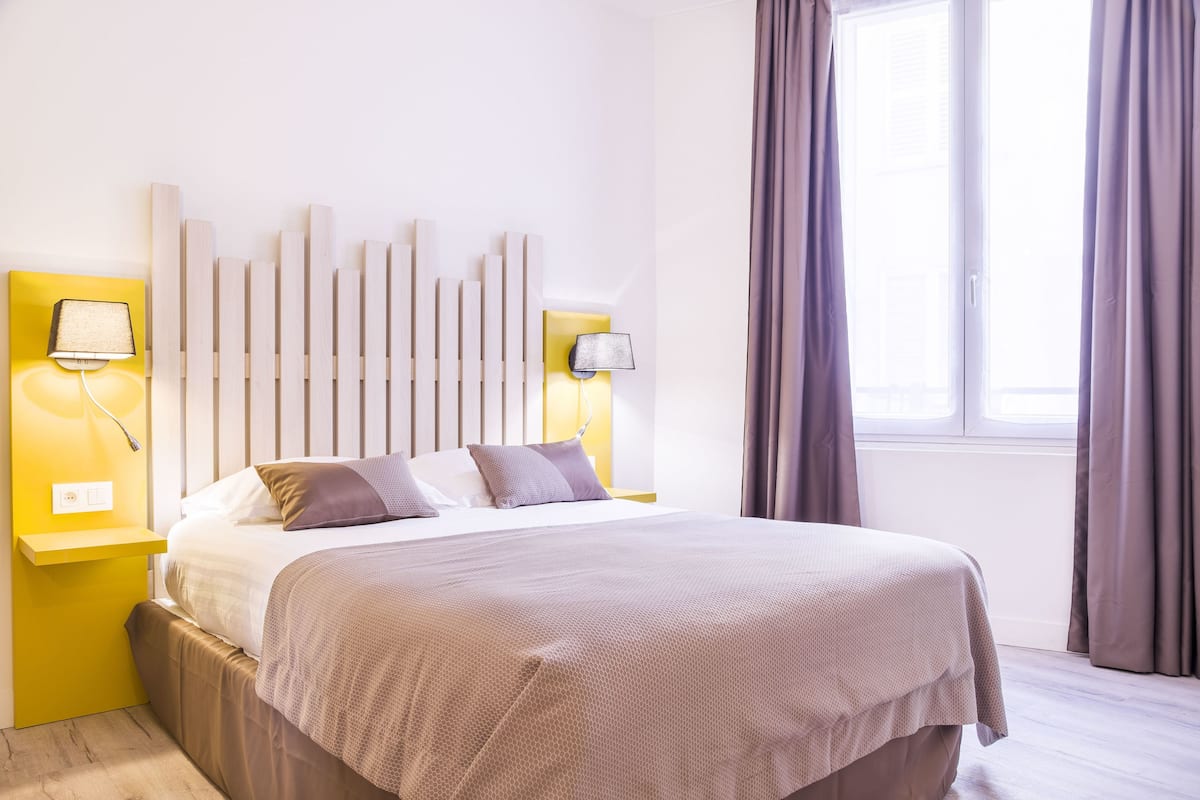 A modern hotel room with a large bed, two bedside lamps, a headboard with vertical slats, and a window with curtains.