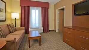 A hotel room with a sofa, a wooden table, a TV, and a window with curtains. - Russell Inn & Suites (Starkville)