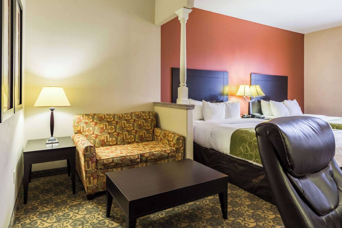 A hotel room with a patterned sofa, a bed with a headboard, a leather chair, a small table, and a lamp.