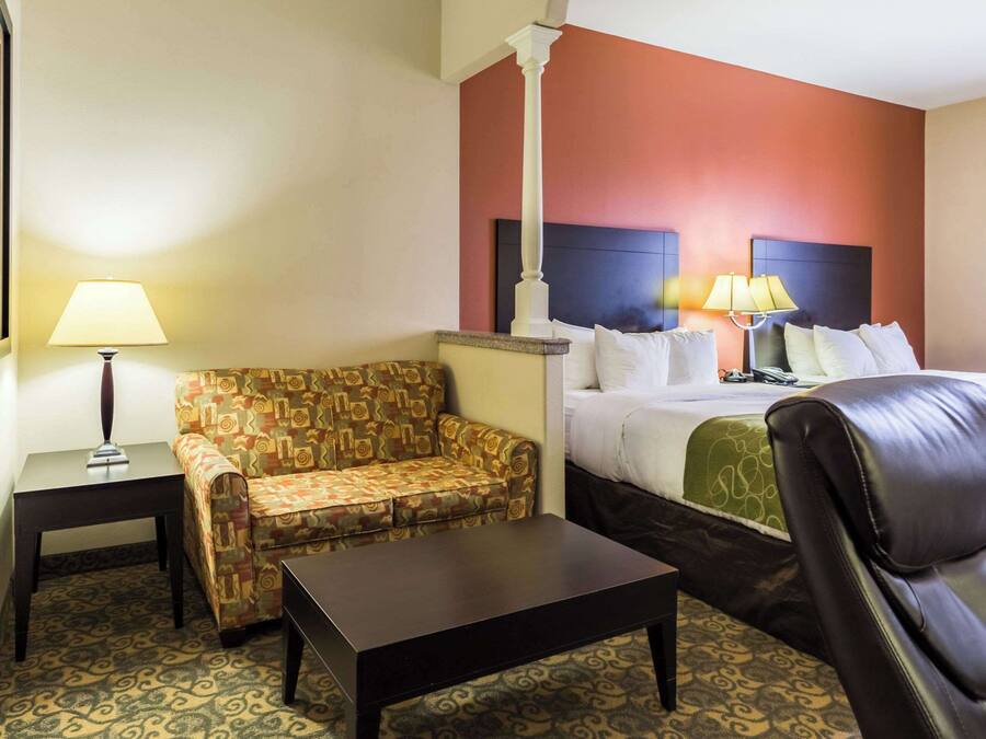 A hotel room with a patterned sofa, a bed with a headboard, a leather chair, a small table, and a lamp.