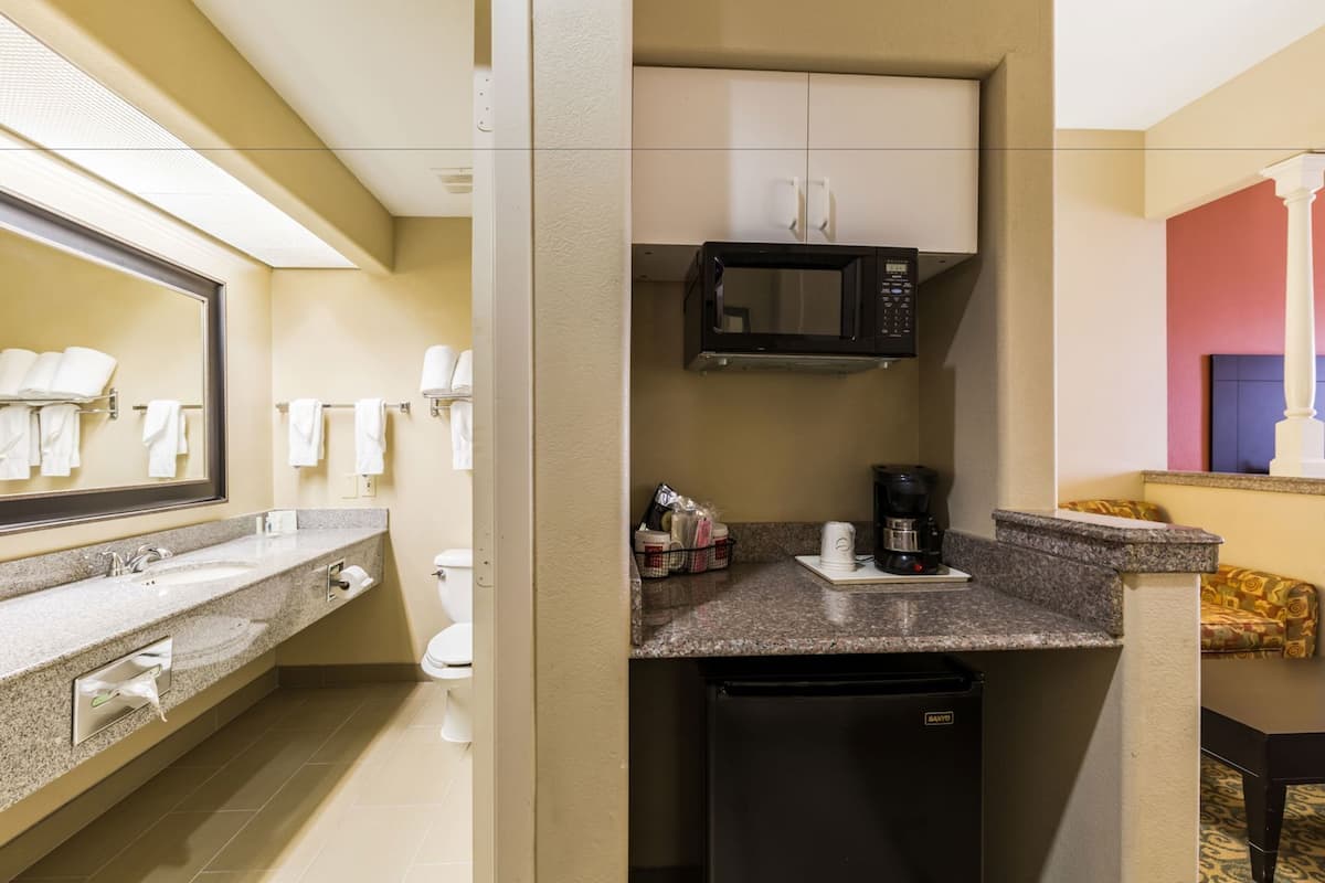 A compact hotel bathroom with a sink, toilet, mirror, microwave, and a small refrigerator.