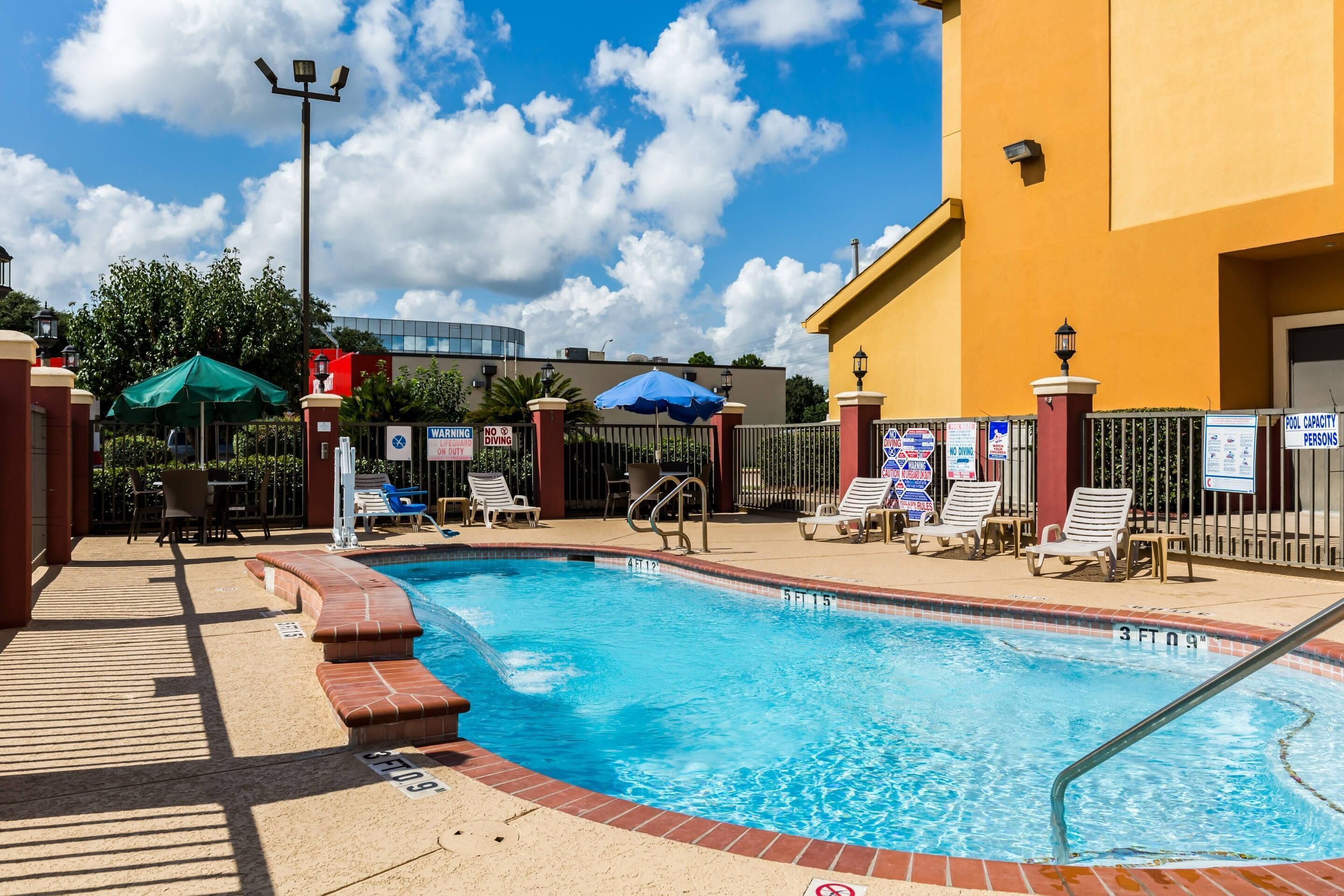 A swimming pool with lounge chairs and umbrellas, surrounded by a fence and a building.