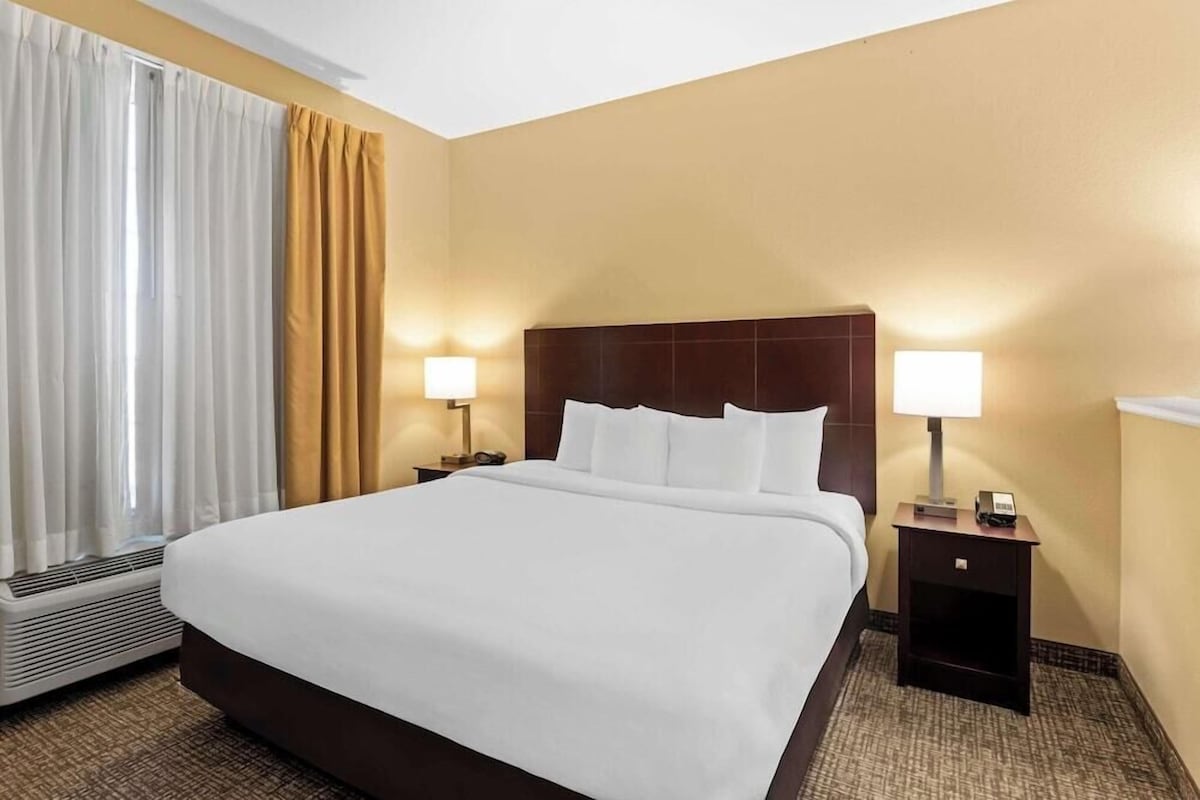 A hotel room with a large bed, two bedside tables with lamps, a window with curtains, and a wall heater.
