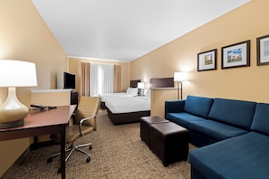 A hotel room with a bed, a desk with a lamp, a blue sofa, a small table, and a TV.